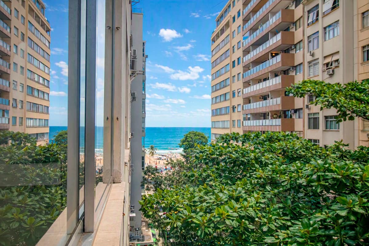 Vista parcial do mar e da praia entre edifícios residenciais, com vegetação em primeiro plano.