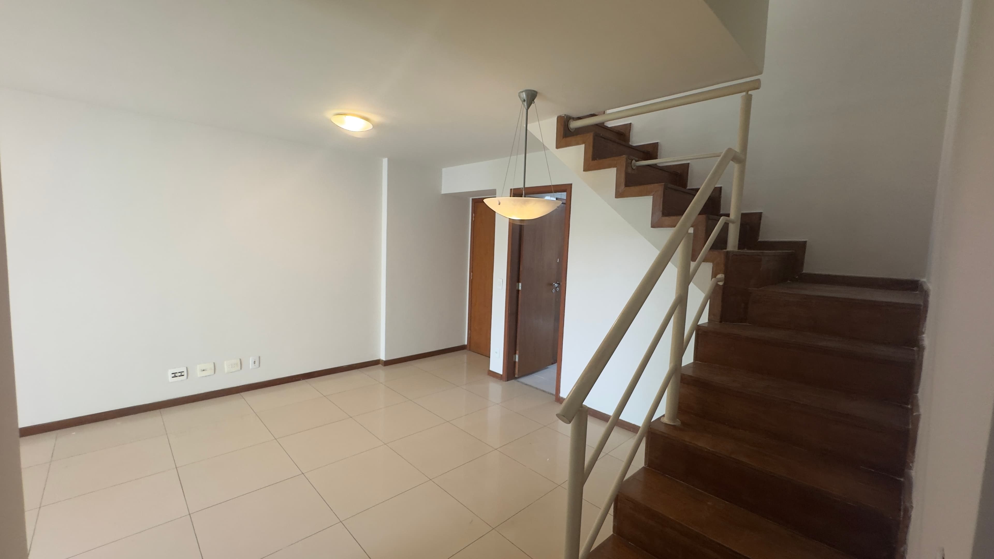 Sala ampla com piso em cerâmica clara, paredes brancas e escada de madeira com corrimão metálico.