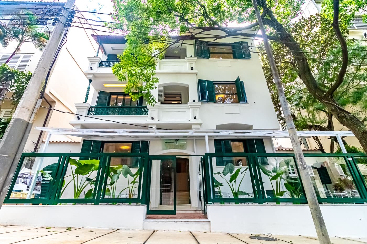 Fachada frontal de prédio branco com detalhes em verde e paisagismo na entrada.